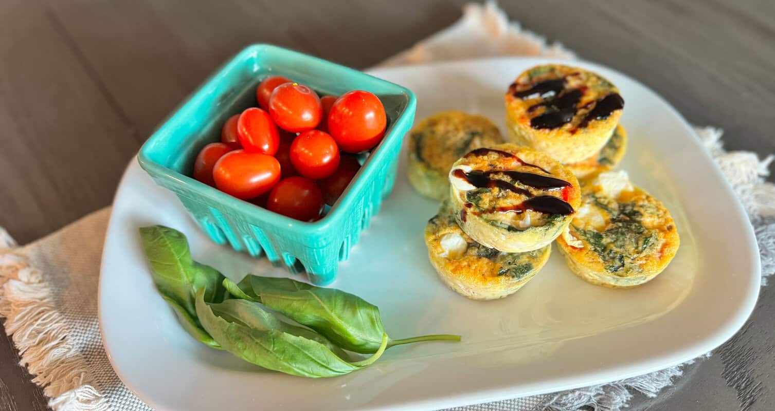 Mini quiches topped with balsamic glaze and basil, served on a white plate with a side of cherry tomatoes in a blue dish.