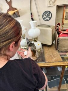 Person looking at a small antique lamp with a price tag in a store, surrounded by vintage items including another lamp, a small box, and a radio.