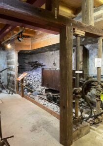 Old mining exhibit with wooden beams, a cart on tracks, rock walls, and mining tools displayed. Lighting illuminates the scene, showcasing a vintage industrial setting.