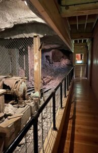Wooden corridor with a railing overlooks a rocky underground mine exhibit. A rusty motor and mining tools are visible behind a wire fence.