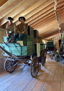 Two mannequins dressed as cowboys sit on a green wooden wagon inside a wooden barn or museum. Other wagons and equipment can be seen in the background.