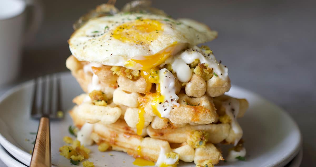 A stack of waffles topped with a fried egg and drizzled with sauce, served on a plate with a fork beside it.