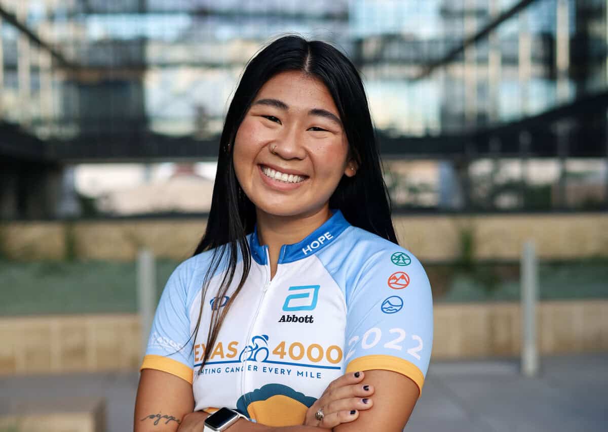 A person smiling and standing outdoors in a cycling jersey with various logos. The background shows a modern building.