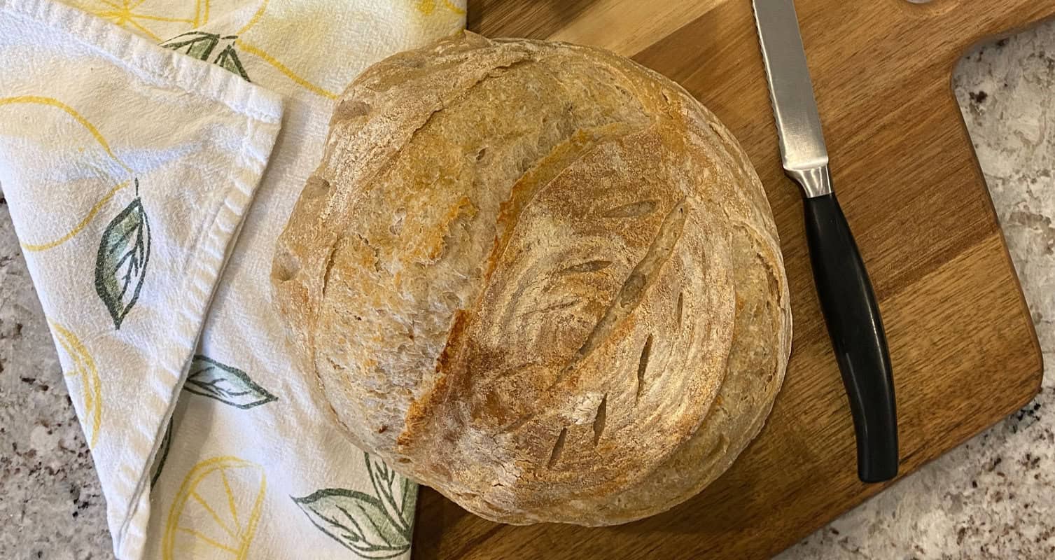 A round loaf of crusty bread on a wooden cutting board beside a knife and a lemon-patterned cloth.