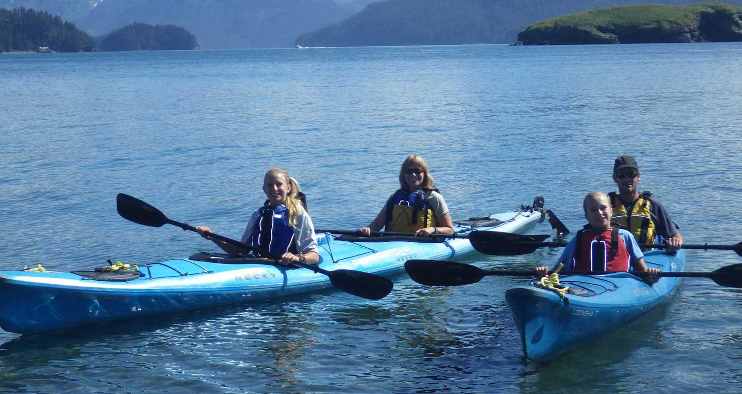 Four people in blue kayaks paddle on a calm lake with a mountainous background.