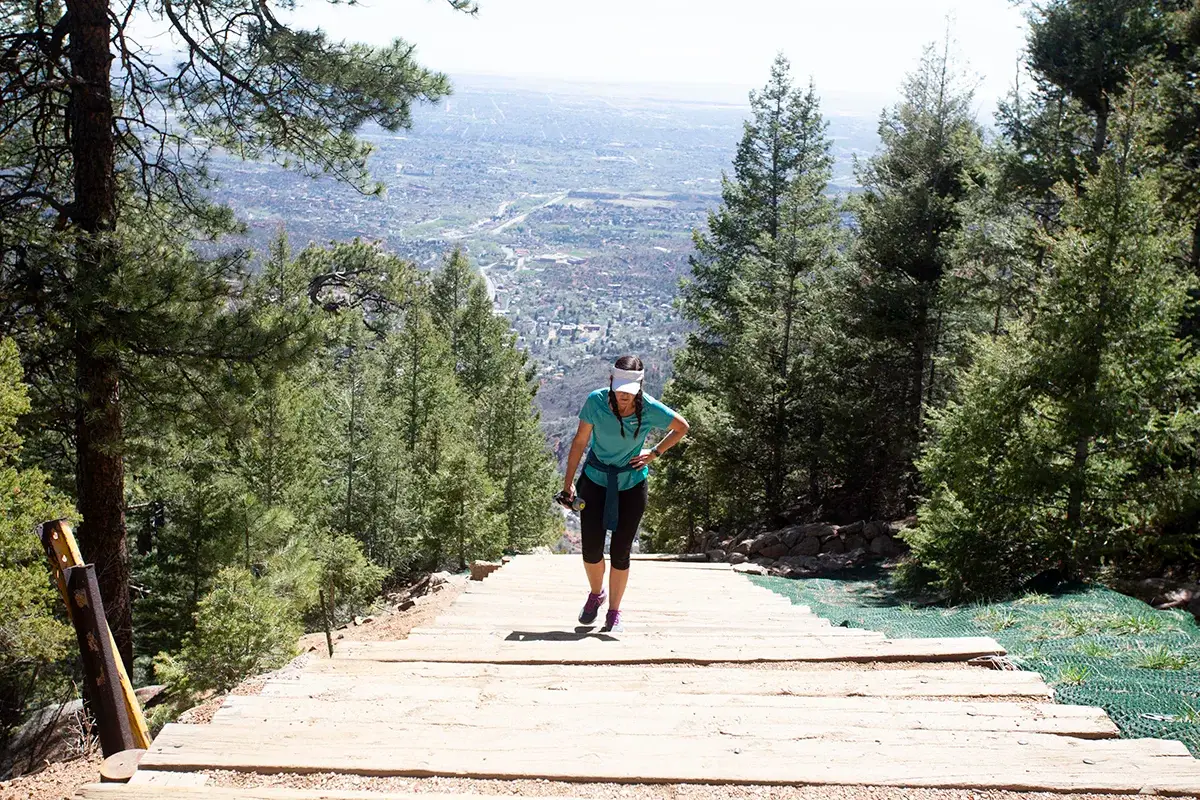 A woman in athletic clothing and a hat climbs wooden steps uphill through a pine forest, with a scenic valley and city visible in the background on a sunny day.