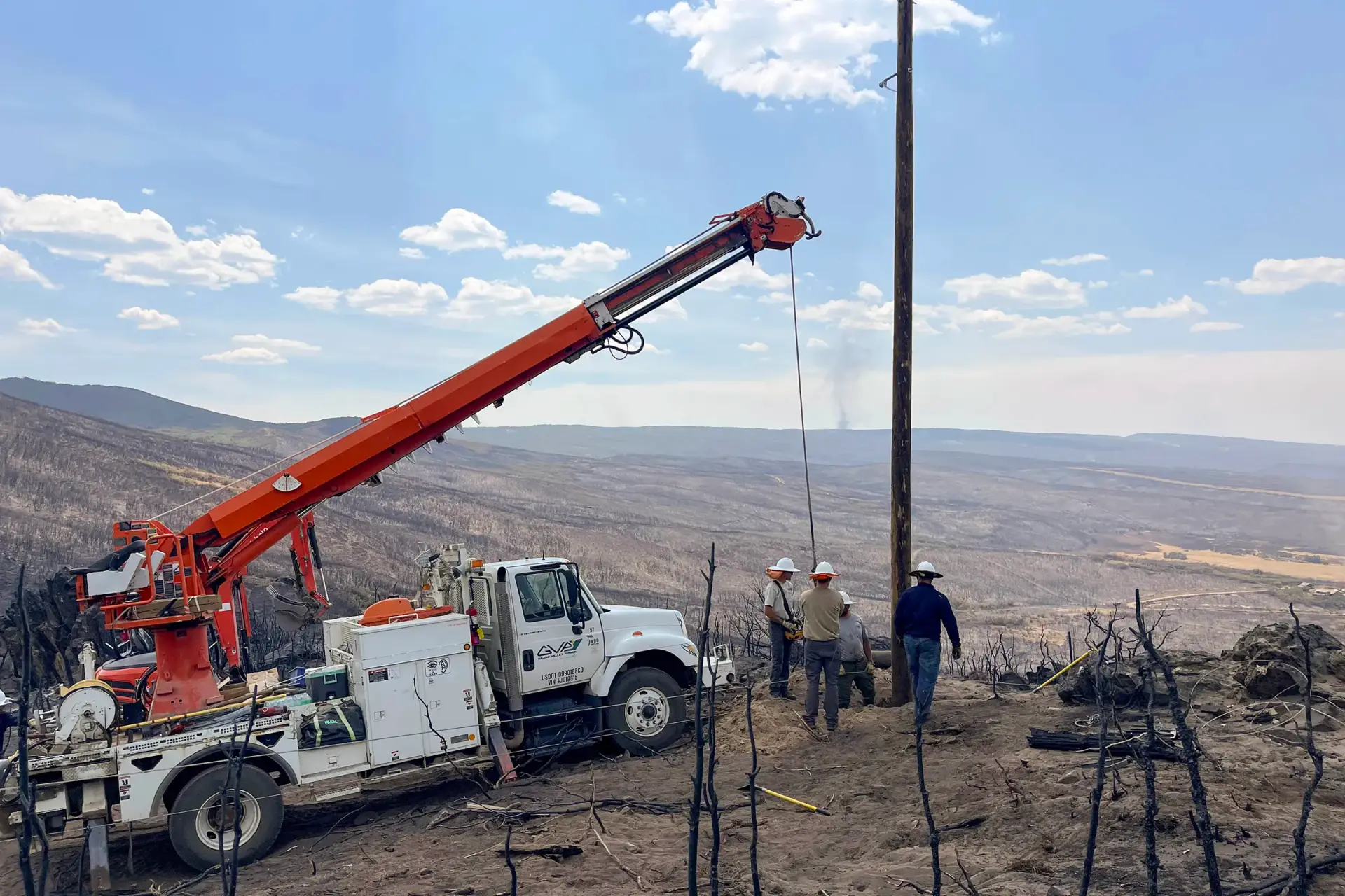 Three workers in hard hats stand near a utility truck with a crane, installing or repairing a utility pole in a rural, fire-damaged landscape.