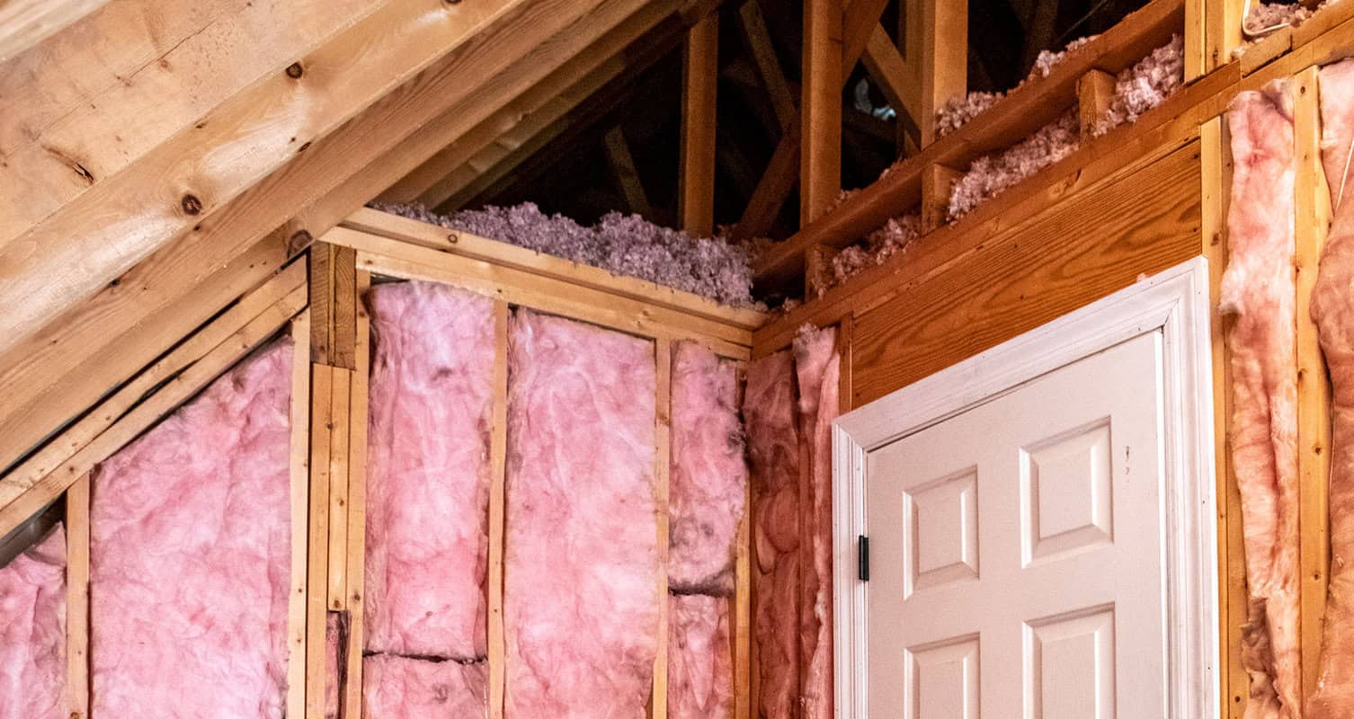 An attic with wooden beams and walls lined with pink fiberglass insulation, next to a closed white door.