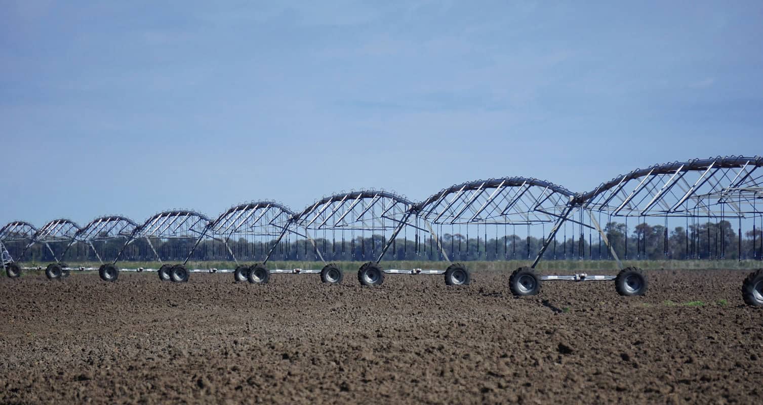 Long row of wheel-mounted irrigation pivots sit idle on a barren field against a blue sky.