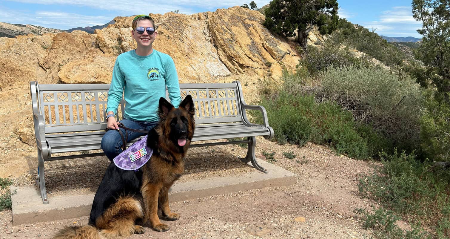 Person sitting on a bench next to a large dog in a mountainous outdoor area.
