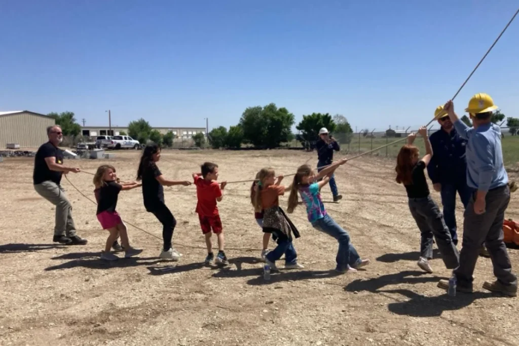 A group of children and adults pull on a rope outdoors in a dirt lot; some adults wear yellow hard hats.
