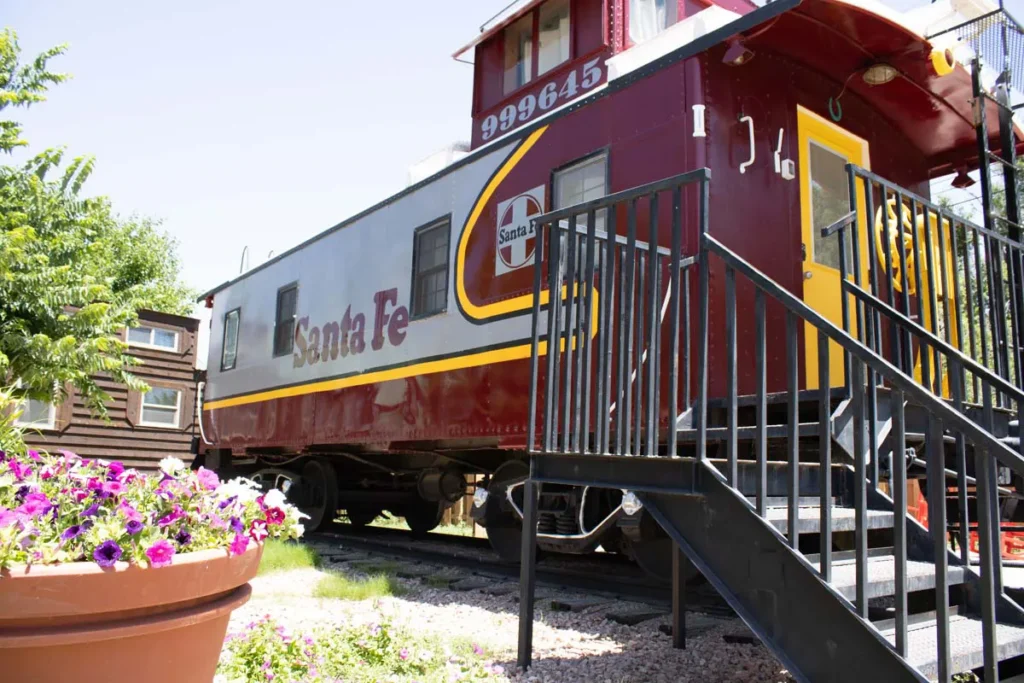 A red and silver Santa Fe caboose with a staircase leading up to its entrance, surrounded by greenery and a pot of colorful flowers in the foreground.