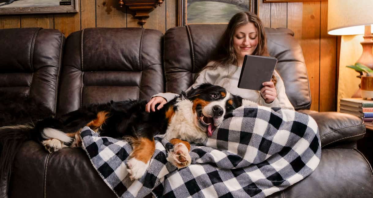 Woman sitting on a couch, reading a tablet, with a large dog lying beside her on a black and white checkered blanket.