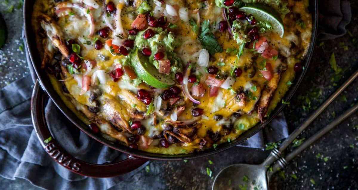 A skillet of cheesy nachos topped with guacamole, diced onions, tomatoes, lime wedges, and pomegranate seeds, with a spoon nearby on a dark surface.
