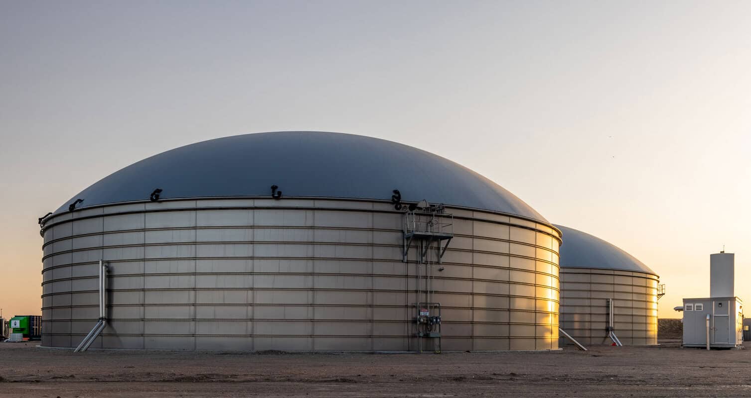 Two large, dome-shaped industrial storage tanks made of metal are situated on a barren landscape under a clear sky at sunset.