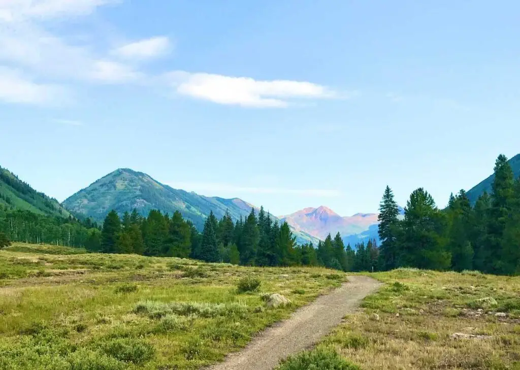 A Short Ride in Co-op Country A dirt path leads through a grassy field with trees, flanked by green mountains under a clear blue sky.