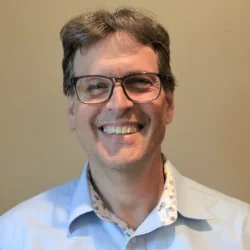 A man with glasses, grayish hair, and a blue collared shirt is smiling at the camera against a plain beige background.
