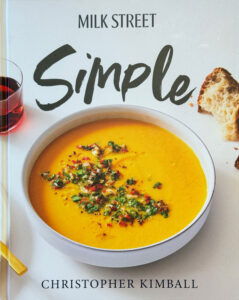 A cookbook titled "Milk Street Simple" by Christopher Kimball, showing a bowl of orange soup garnished with herbs, next to bread and a glass of red liquid.