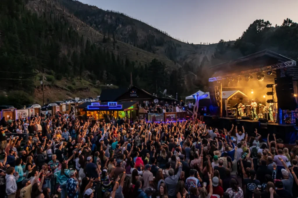 A large crowd attends an outdoor concert at dusk in a mountainous area, with a lit stage and food stands visible in the background.