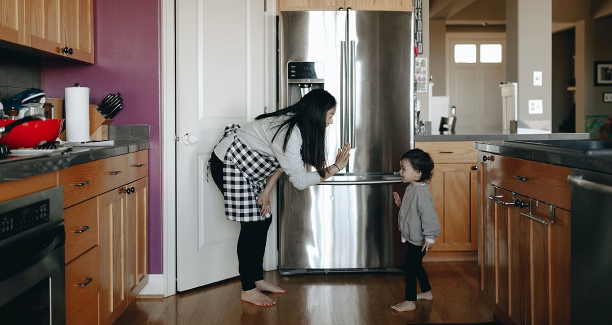 Woman bending to talk to a toddler in a kitchen with wooden cabinets and a stainless steel refrigerator.