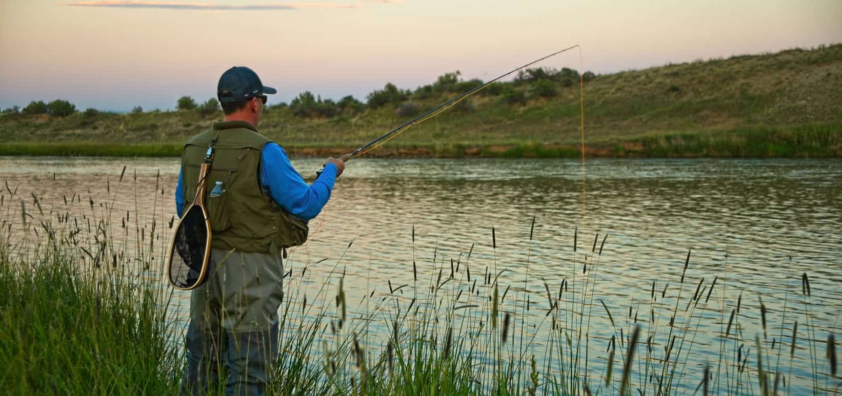 A person wearing fishing gear is standing on grassy riverbank, holding a fishing rod over a calm river at sunset.