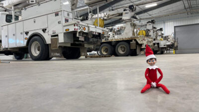 Elf doll sitting on the floor in an industrial garage with several large utility trucks in the background.
