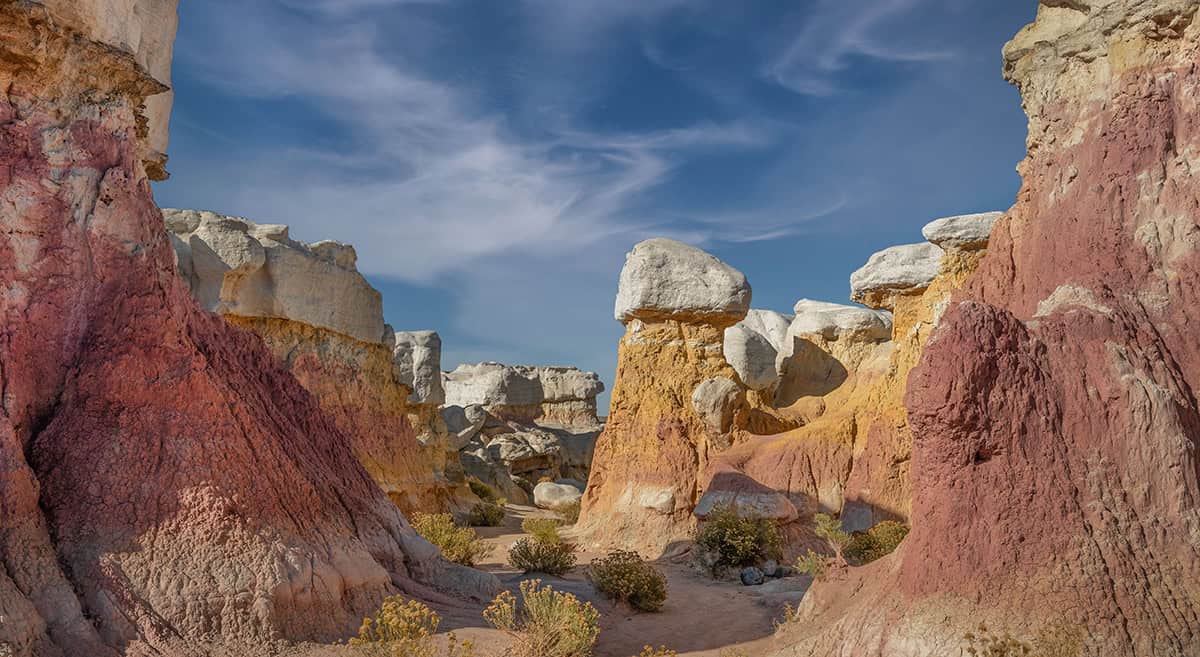 Rock formations with red, orange, and white layers under a blue sky with wispy clouds. Sparse vegetation is visible at the base.