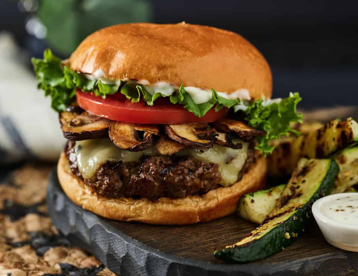 Cheeseburger with grilled mushrooms, lettuce, tomato, and mayonnaise on a bun, served with roasted zucchini and a side of sauce.