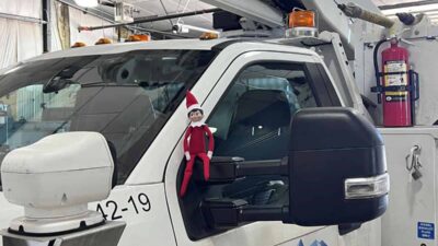 A small toy elf is sitting on the driver's side mirror of a utility truck inside a garage. The truck has a large boom arm and various company logos on the door.