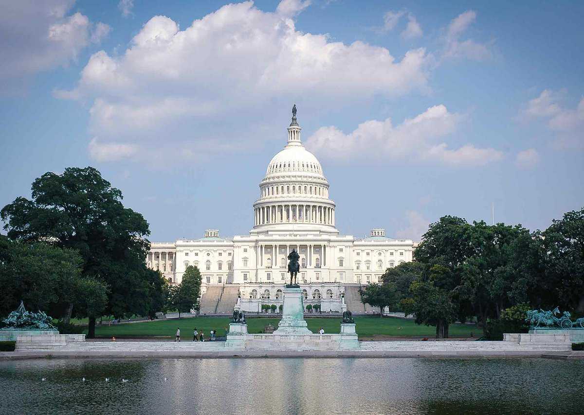The image shows the U.S. Capitol building with its iconic dome, surrounded by green lawns and trees, under a partly cloudy sky. A fountain is visible in the foreground.