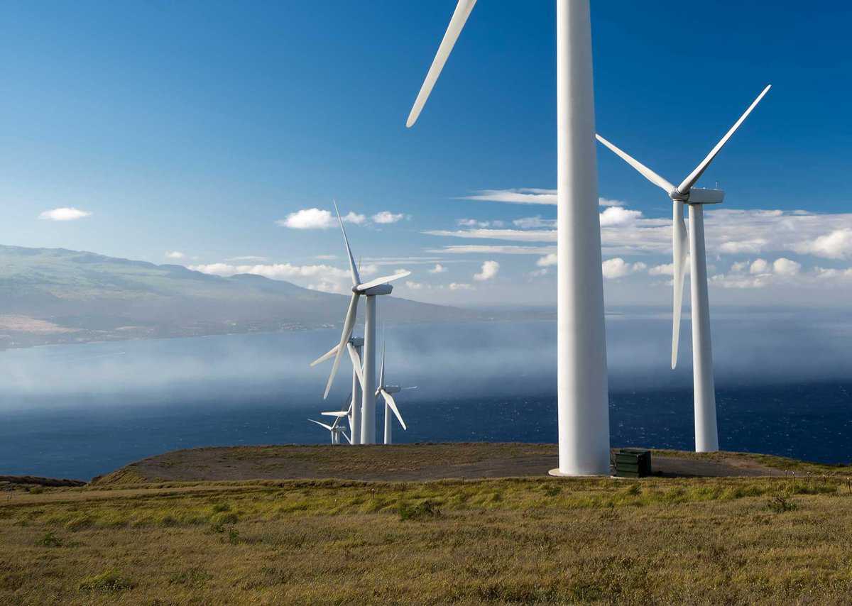 Wind turbines situated on a grassy hill with a vast ocean in the background under a clear blue sky.