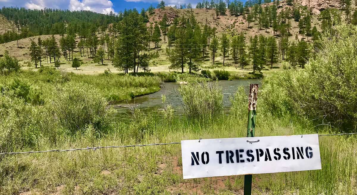 Sign reads "No Trespassing" on a fence in front of a grassy landscape with trees, hills, and a river under a blue sky.