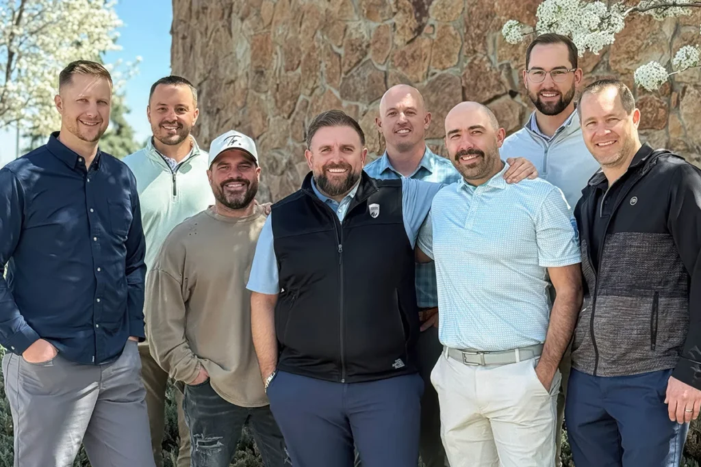 Eight men stand together outdoors in front of a stone wall, posing for a group photo on a sunny day.