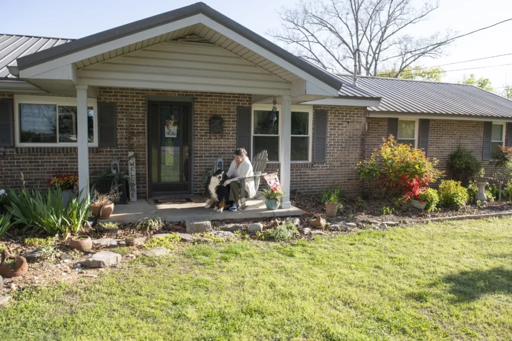 A person sits with a dog on the front porch of a single-story brick house with a well-maintained garden and lawn.