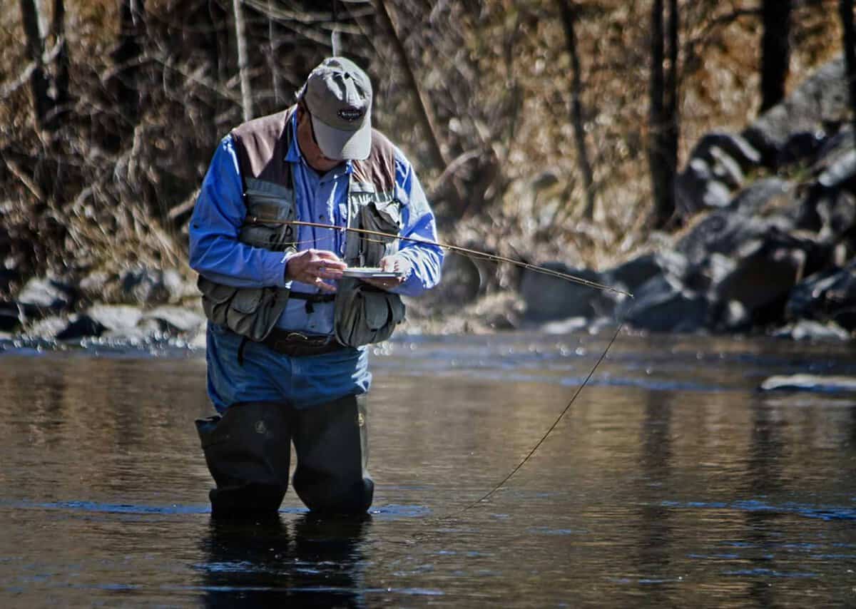 Person standing in a river, wearing a cap and fishing vest, looking down while holding fishing gear.