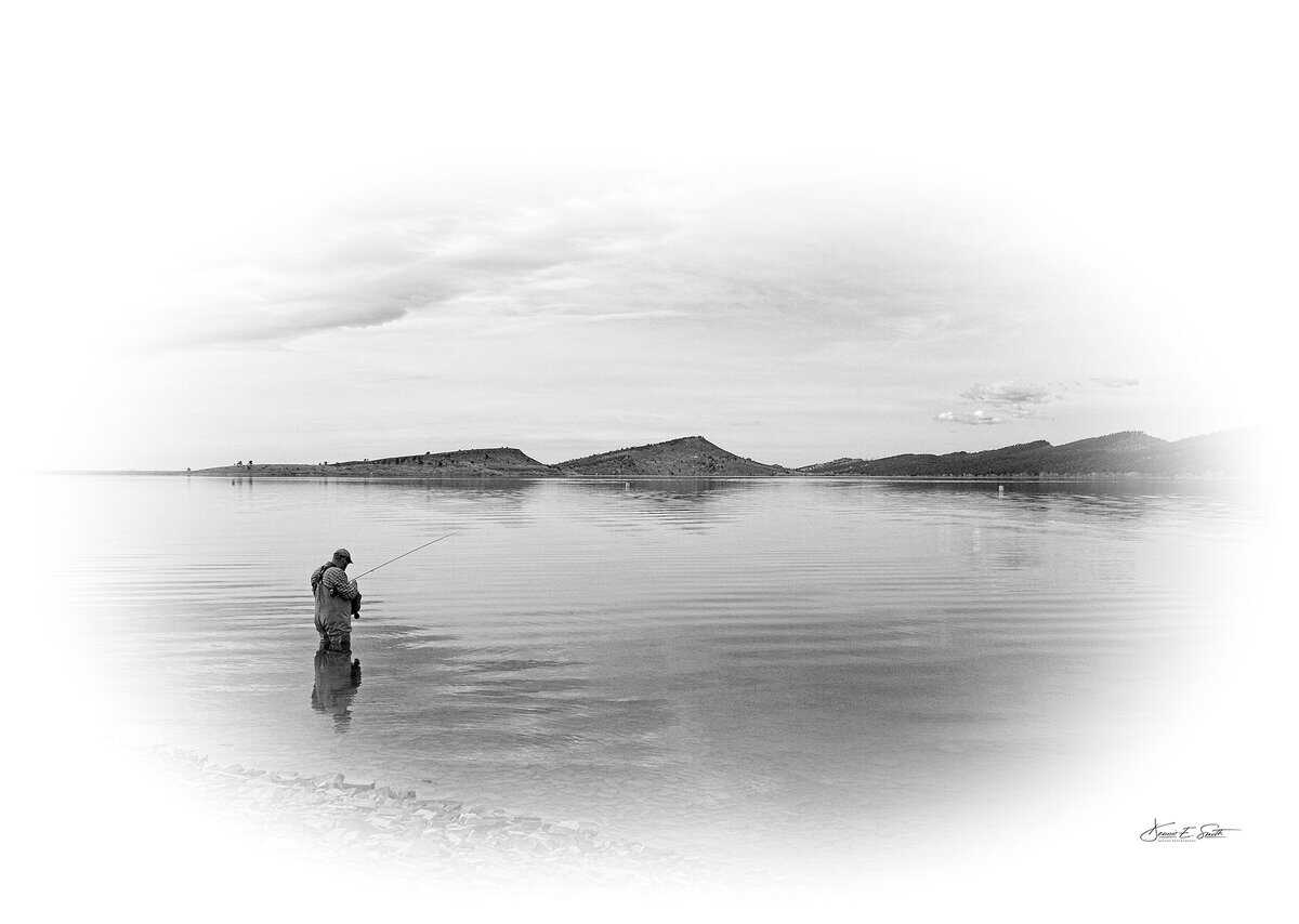 A person stands knee-deep in calm water while fishing, with distant hills under a cloudy sky. Black and white image.