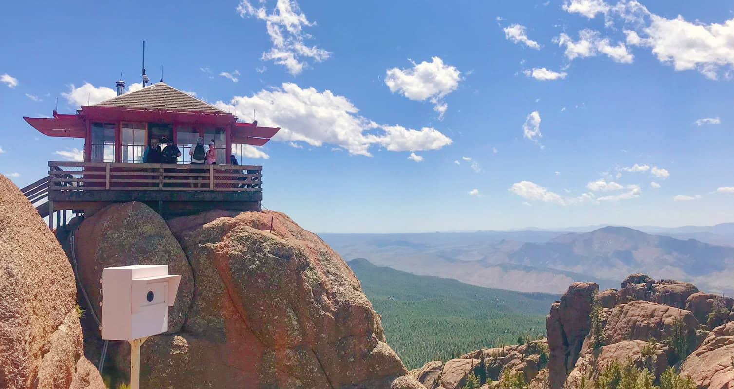 A wooden lookout station perched on a rocky cliff overlooks a vast forest and mountain range under a clear blue sky with scattered clouds.