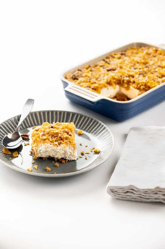 A square piece of creamy dessert with a crumbly topping on a plate with a spoon, next to a baking dish containing more of the dessert and a folded napkin.