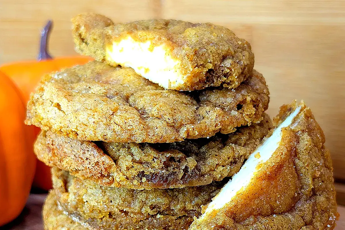 A stack of spiced cookies with creamy white filling, set against a wooden background with a small orange pumpkin nearby.