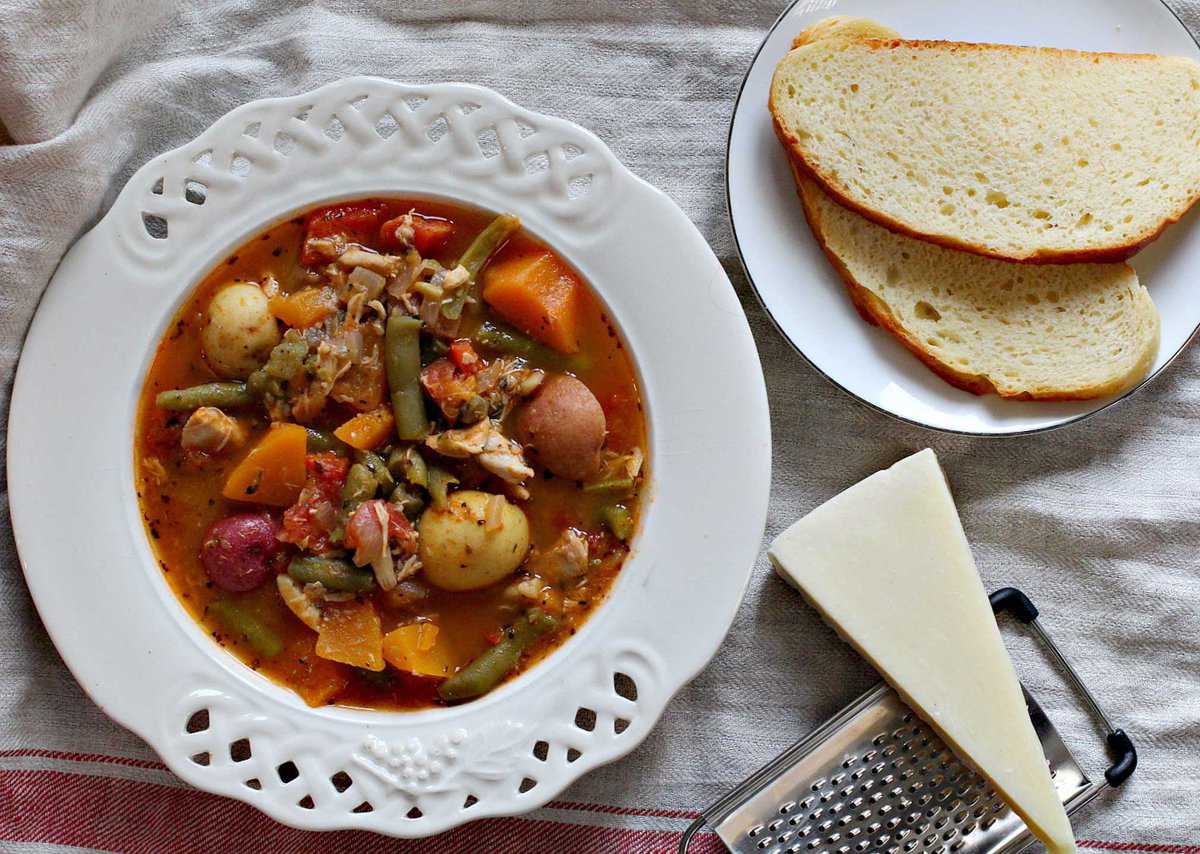 A bowl of vegetable and chicken soup with potatoes, carrots, and green beans on a lace-edged plate. A side of sliced bread and a wedge of cheese are nearby on a tablecloth.