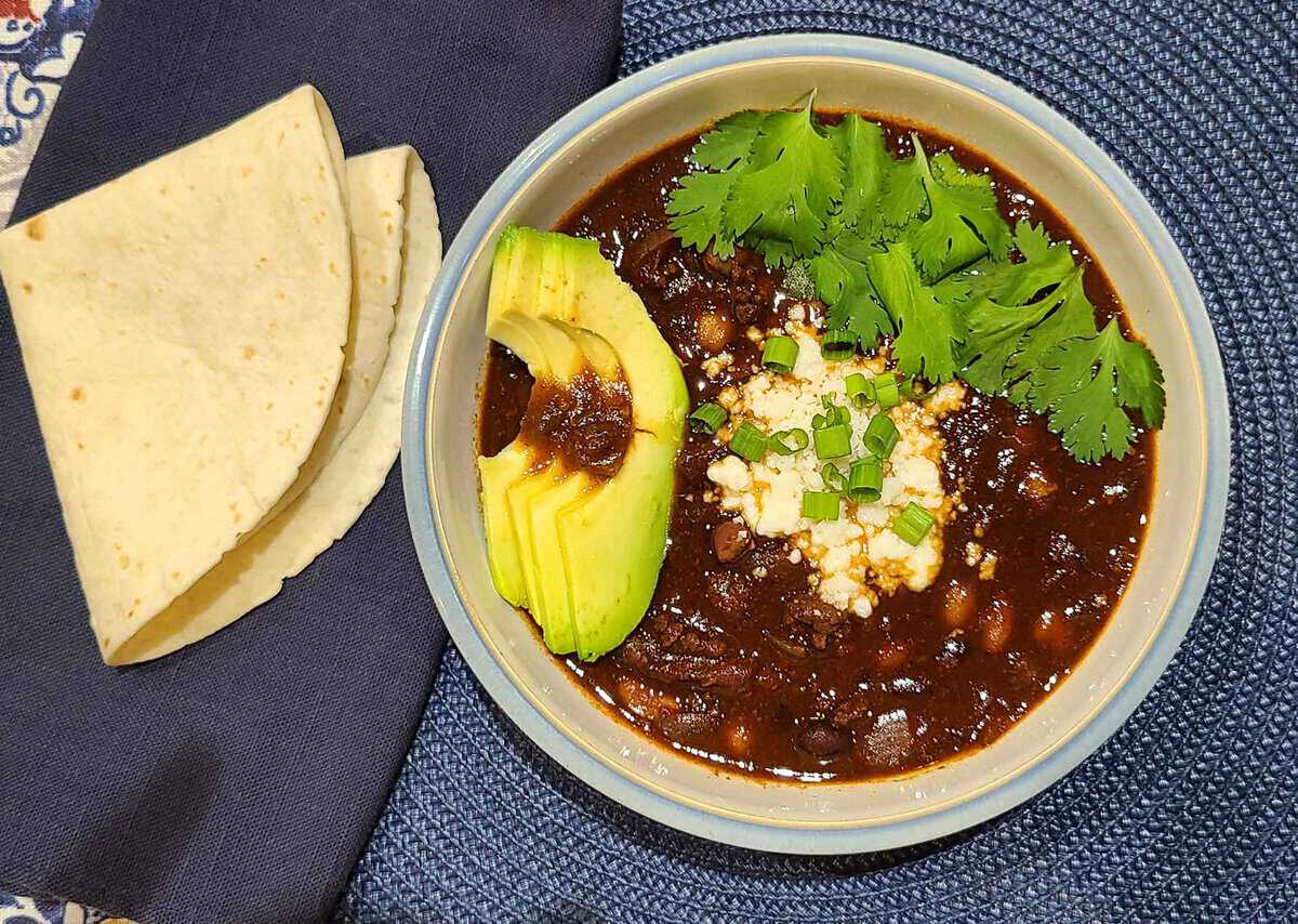 Bowl of black bean soup garnished with avocado slices, cilantro, cheese, and chives, next to folded tortillas on a dark cloth.