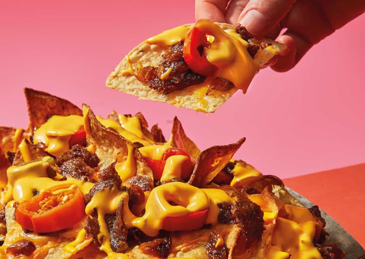 A hand lifting a nacho chip topped with melted cheese, jalapeños, and beef from a plate filled with similar nachos; pink background.
