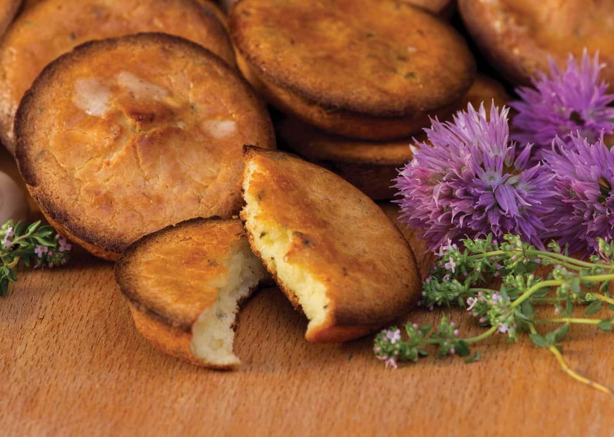 Golden-brown biscuits with one partially eaten, surrounded by purple flowers and green herbs on a wooden surface.