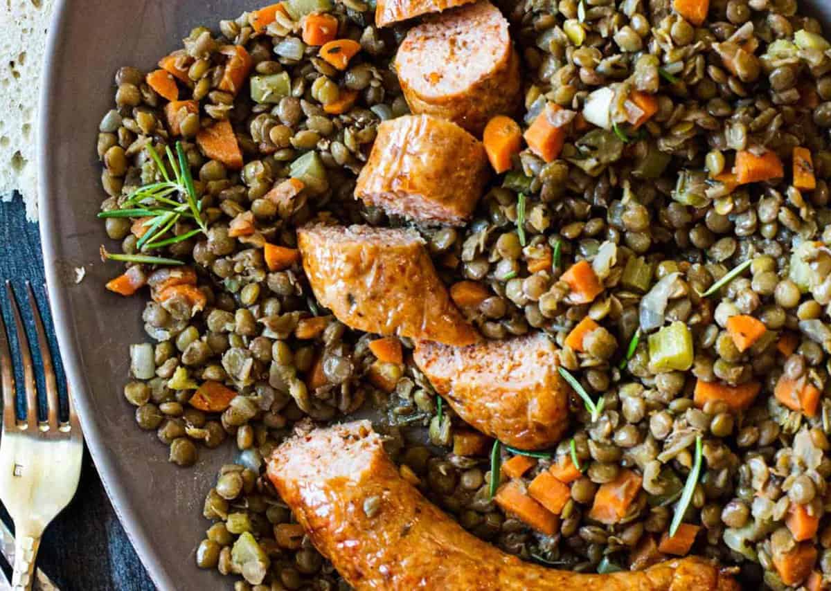 Plate of cooked lentils with carrots, celery, and rosemary, topped with sliced sausage. Fork on the side.
