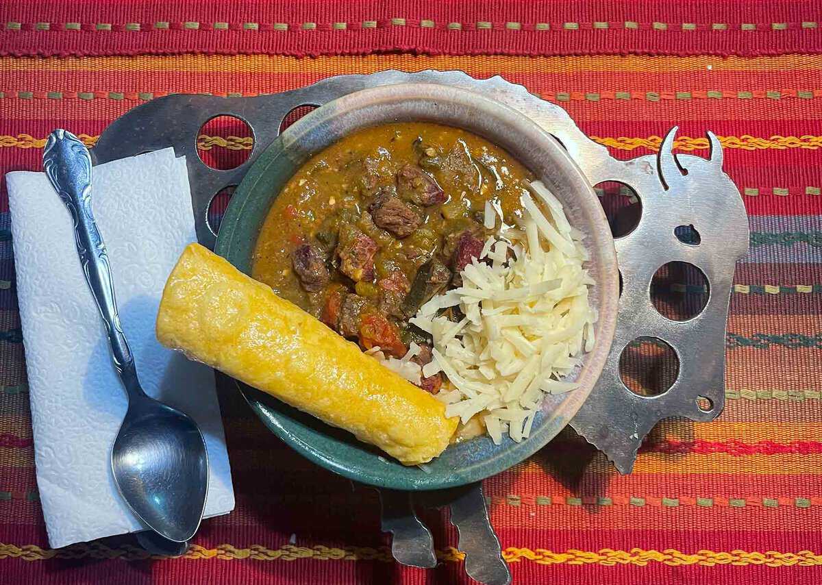 Bowl of stew with shredded cheese and a rolled tortilla, placed on a metal trivet decorated with a cow design, next to a spoon on a napkin, over a woven red placemat.