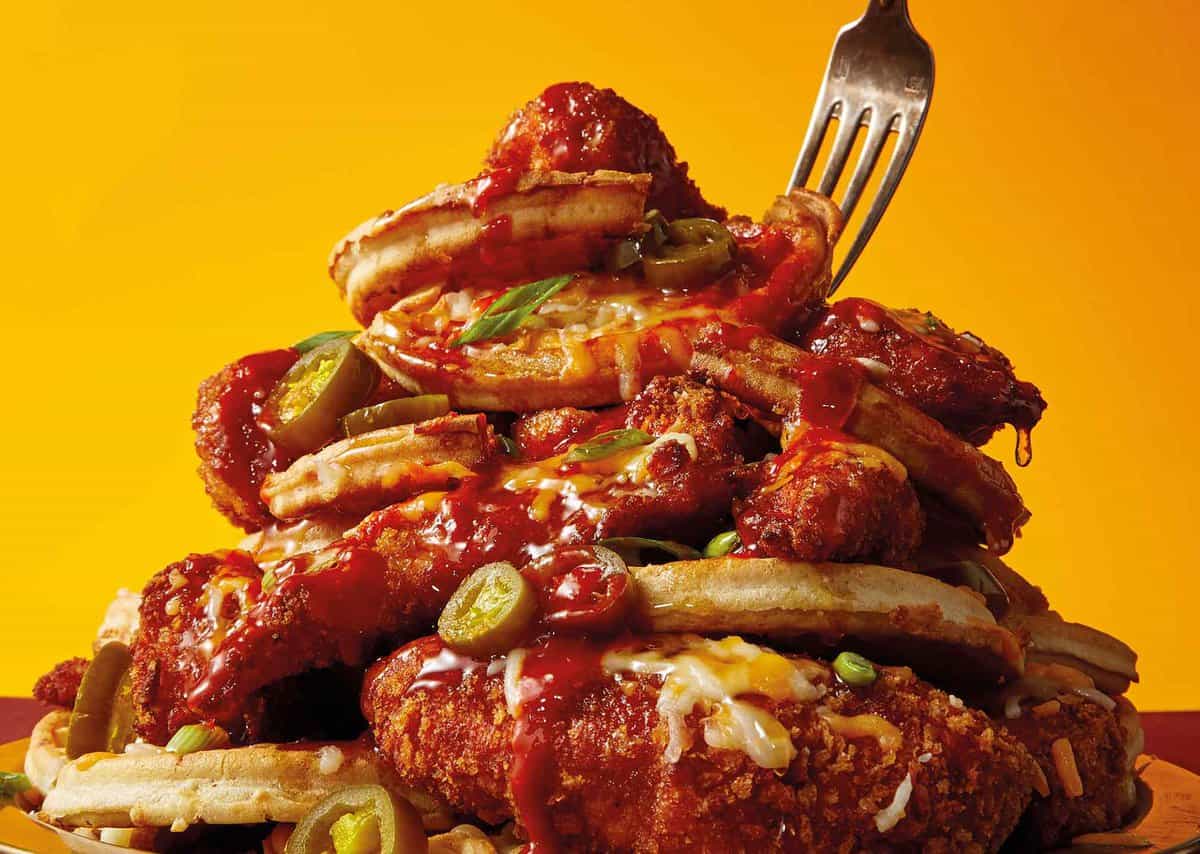 A stack of waffles and fried chicken, garnished with jalapeños, green onions, cheese, and drizzled with sauce. A fork is inserted into the top.