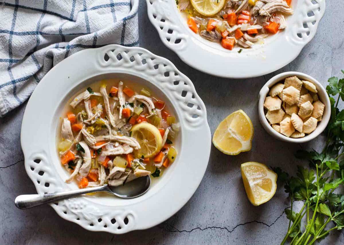 Two white bowls of chicken soup with vegetables and lemon slices on a table, accompanied by lemon wedges, oyster crackers, and a cloth napkin.