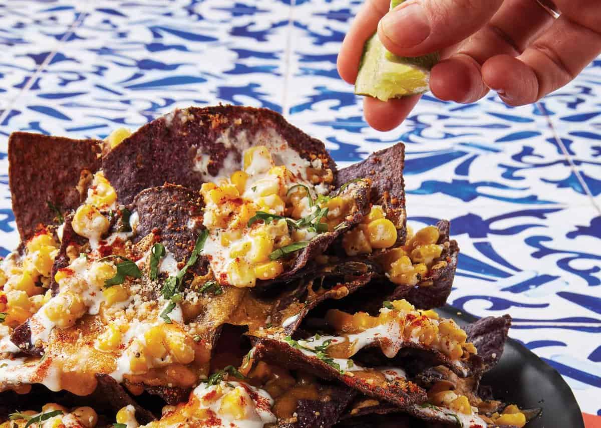 A hand squeezes lime over blue corn nachos topped with melted cheese, corn, herbs, and spices on a patterned blue and white table.