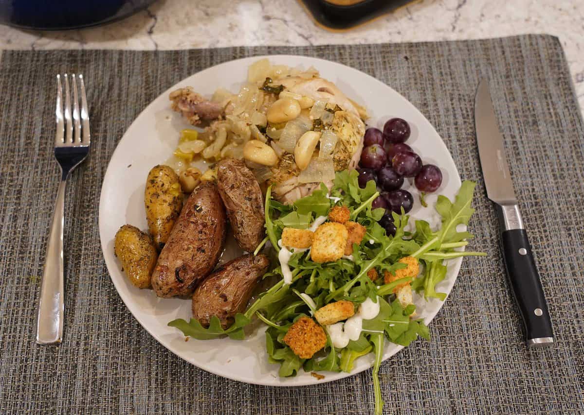 A plate with roasted chicken, potatoes, salad with croutons and dressing, and a small bunch of grapes, set on a placemat with a fork and knife beside it.