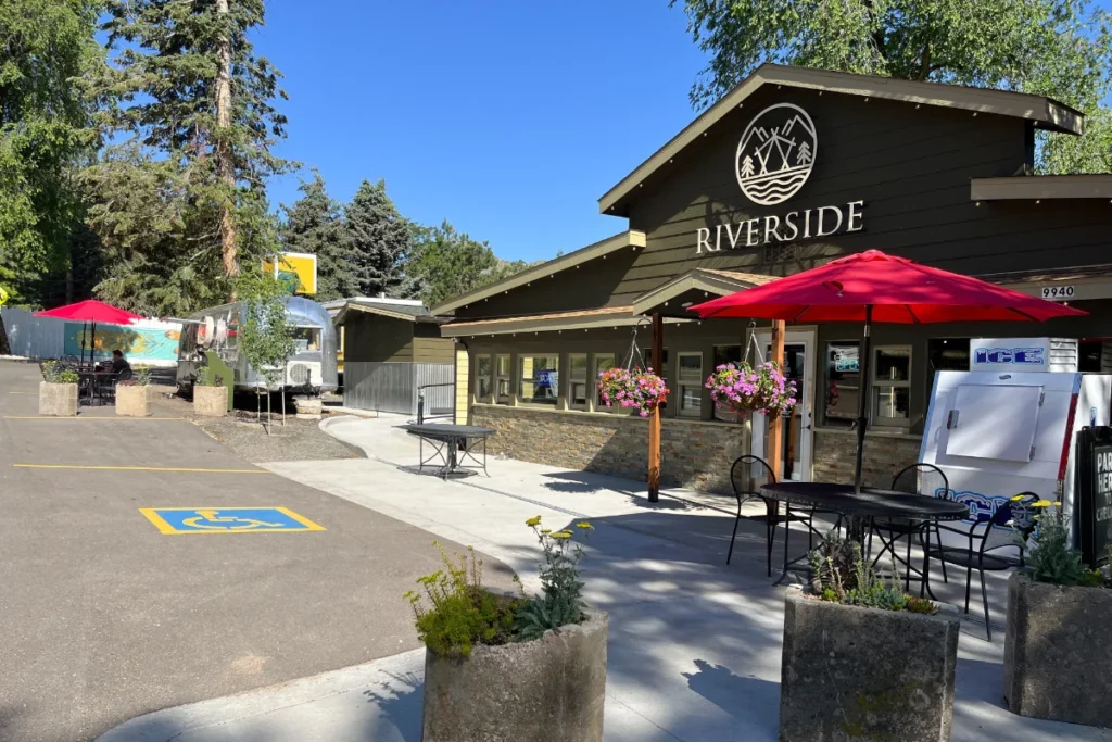 Outdoor seating area with red umbrellas in front of a building labeled “Riverside,” next to an ice machine, planters, and a marked accessible parking space.